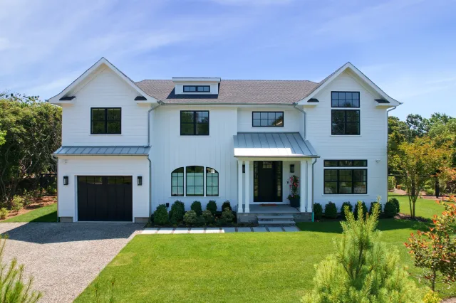 Modern two-story white farmhouse with black windows, a garage, and a large green lawn under a clear blue sky