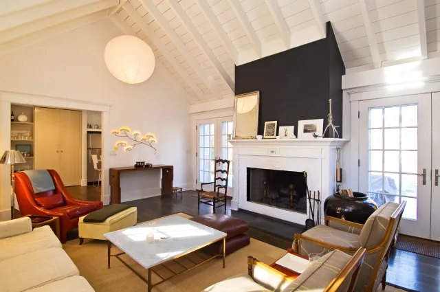 Bright living room with white vaulted ceiling, fireplace, leather and wooden chairs, and large windows.