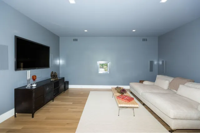 Modern living room with light gray couch, wooden floor, wall-mounted TV, and small window on light blue walls.