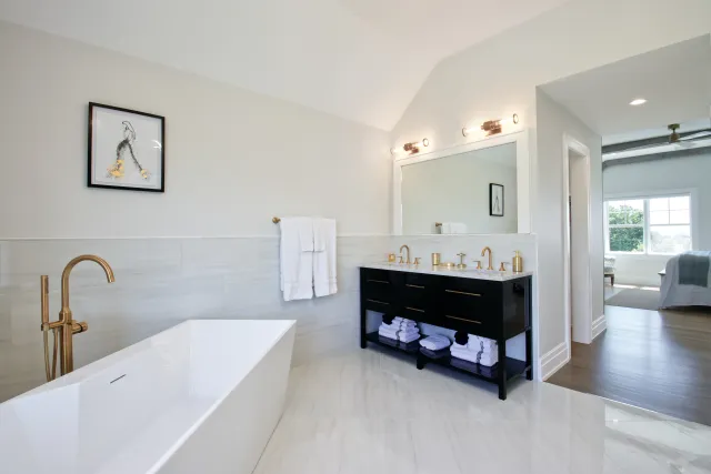 Modern bathroom with a white freestanding tub, black vanity with gold fixtures, and large mirror under soft lighting.