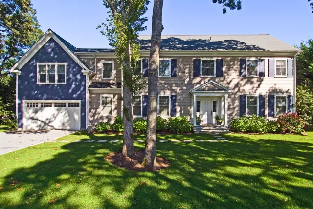 Two-story suburban house with gray and blue siding, white trim, black shutters, and a green lawn under clear sky.