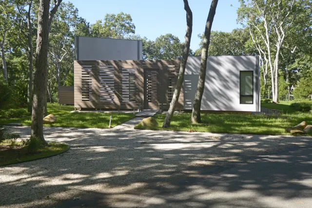 Modern single-story house with wooden and white exterior surrounded by trees and green lawn under sunlight.
