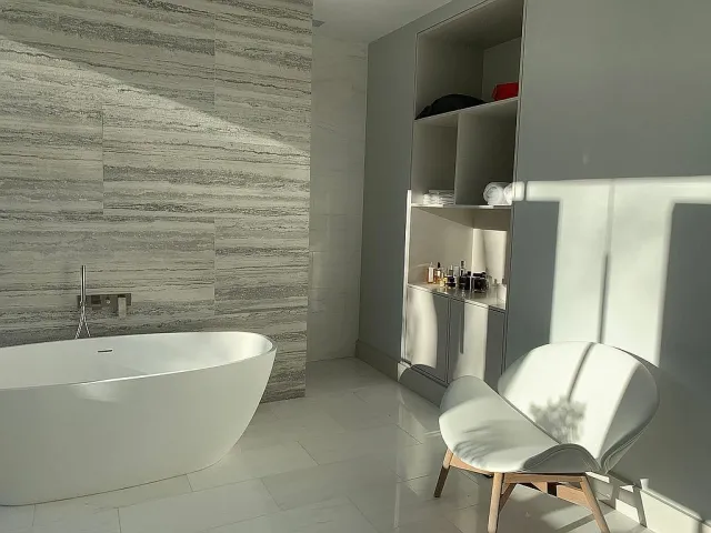 Modern bathroom with freestanding white tub, textured wall, built-in shelves, and white accent chair with sunlight.