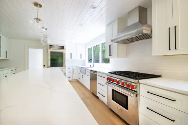 Bright modern kitchen with white cabinetry, marble countertops, stainless steel appliances, and large windows.