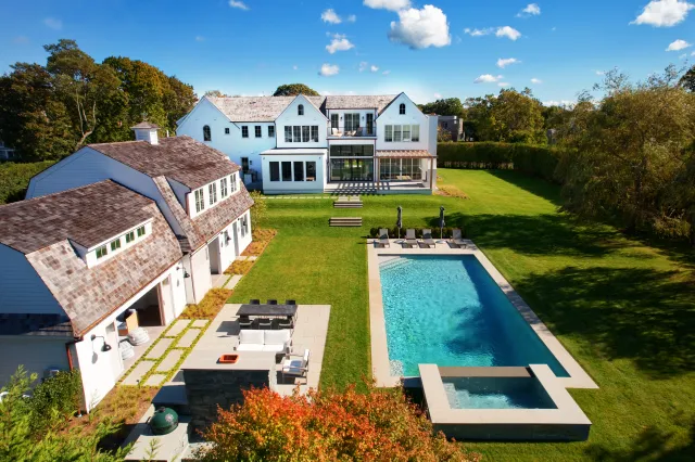 Modern white house with large backyard, rectangular swimming pool, patio seating, and lush green lawn on sunny day