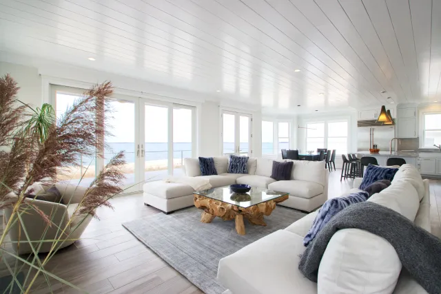 Bright modern living room with white sectional sofa, glass coffee table, ocean view, and wooden flooring.