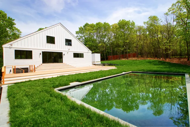 Modern white house with large wooden deck adjacent to a reflective backyard pond surrounded by green lawn and trees