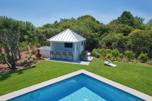 Backyard with rectangular pool, white poolside bar, bar stools, grill, and grass surrounded by trees under clear sky