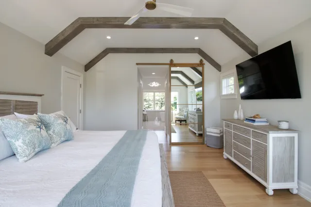 Bright modern bedroom with rustic wooden beams, white walls, light wood flooring, and a large TV on the wall.