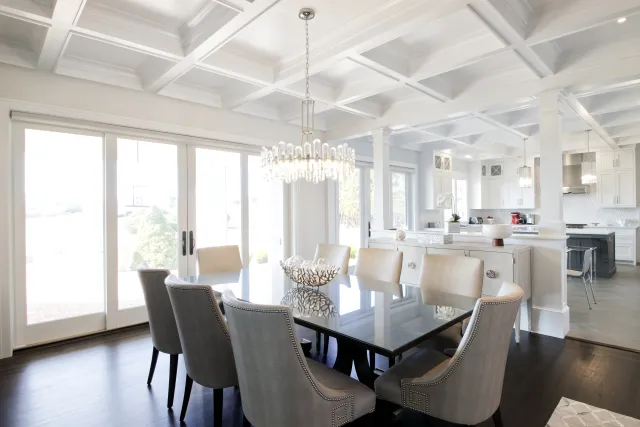 Bright modern dining room with glass table, upholstered chairs, crystal chandelier, and open white kitchen.