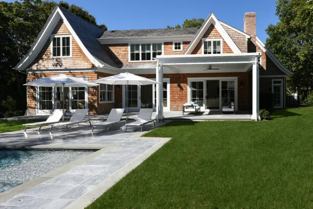 Spacious backyard with a modern house, swimming pool, lounge chairs, umbrellas, patio, and lush green lawn on a sunny day.
