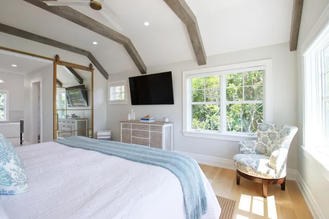 Bright bedroom with vaulted ceiling, wood beams, large window, floral chair, and wall-mounted TV over dresser.