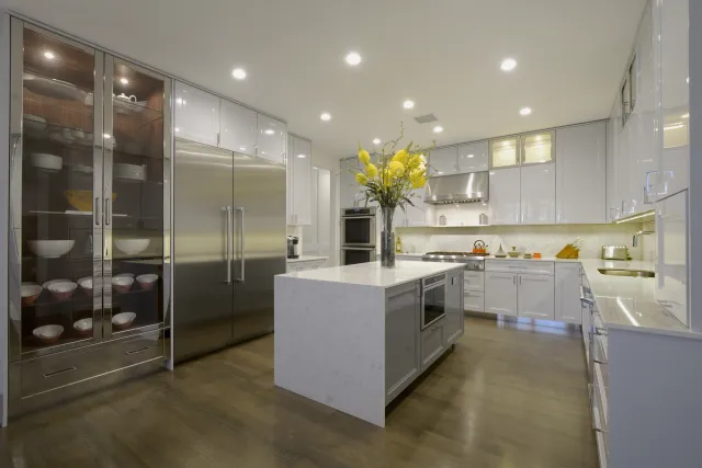 Modern white kitchen with large island, stainless steel appliances, glass cabinet, and hardwood flooring under bright lights