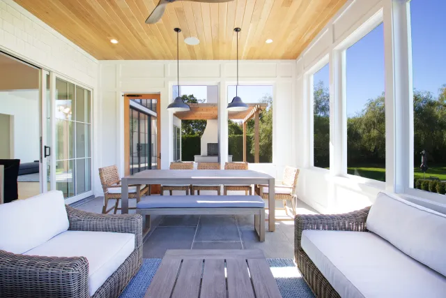 Bright modern sunroom with wicker furniture, wooden ceiling, large windows, and a dining area with pendant lights.