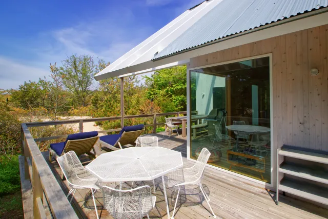 Outdoor wooden deck with white dining table, chairs, lounge chairs, and greenery under blue sky.