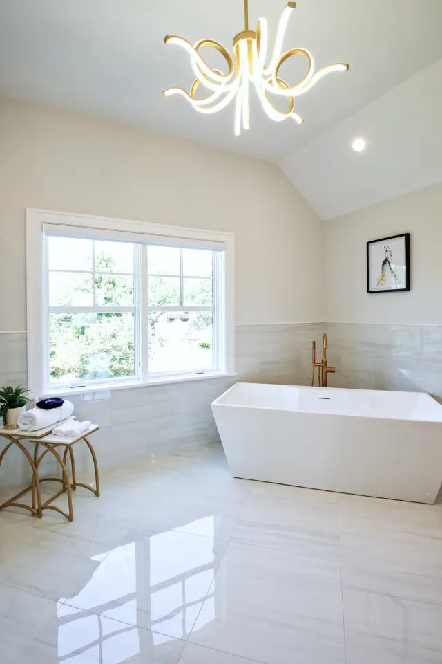 Bright modern bathroom with a freestanding white bathtub, large window, and stylish chandelier.