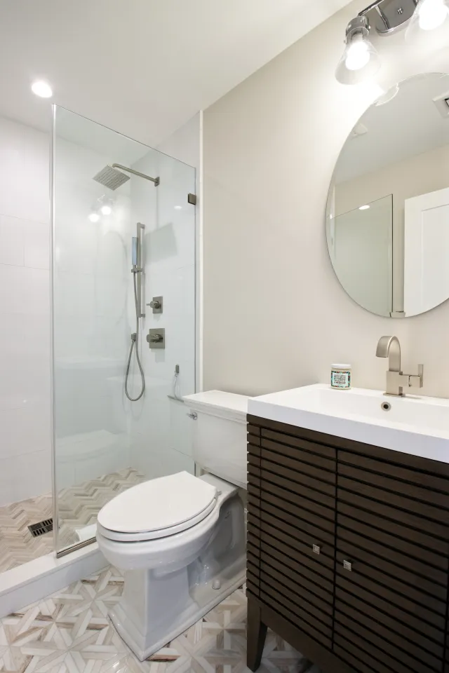 Modern bathroom with glass shower enclosure, white toilet, wood vanity with white sink, and round mirror.