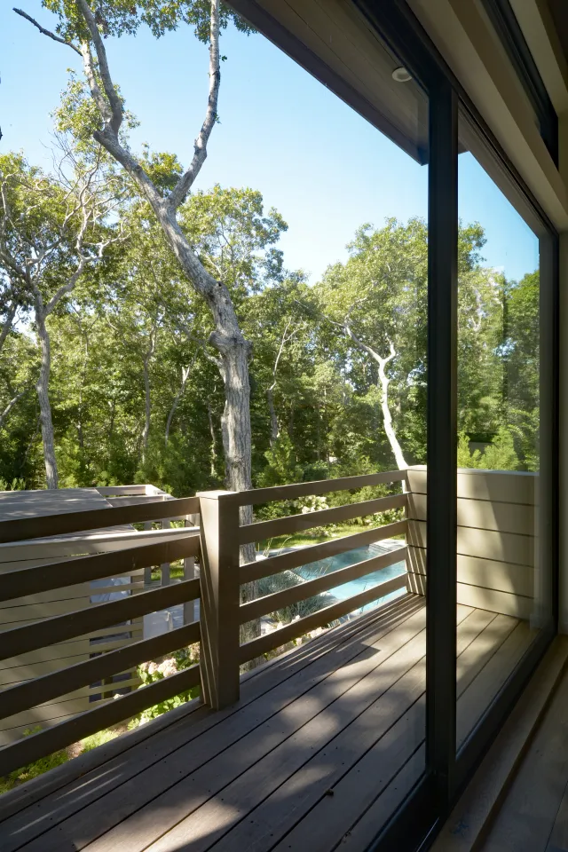 Wooden balcony with railing overlooking a lush green forest under clear blue sky through glass doors