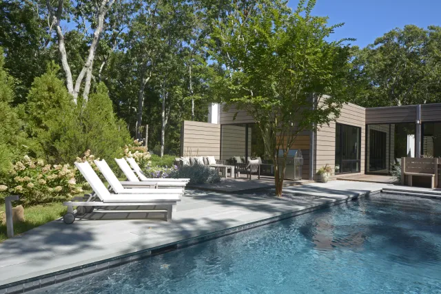 Modern backyard with a clear swimming pool, white loungers, trees, and a contemporary home under a blue sky.