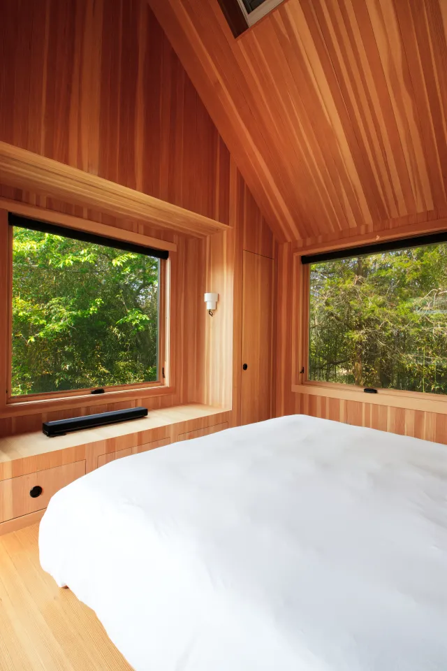 Bright bedroom with wood-paneled walls, large windows showing green trees, and a white bedspread.