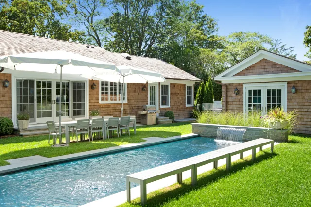 Backyard with modern narrow pool, wooden bench, dining table with umbrellas, and two shingle-style houses.