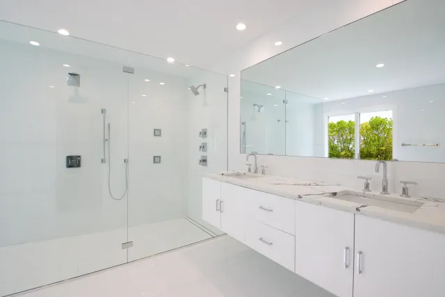 Bright modern white bathroom with glass shower enclosure, double sinks, large mirror, and greenery outside window