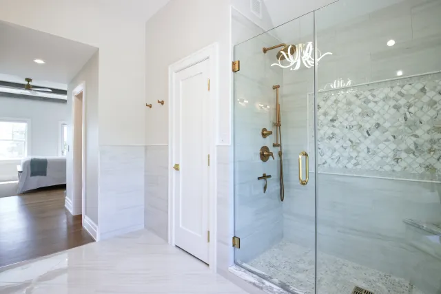 Modern bathroom with glass shower enclosure, gold fixtures, marble tiles, and a view into a bedroom with hardwood floor.