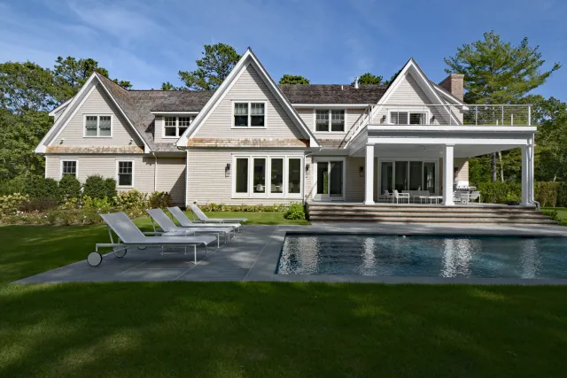 Large modern house with multiple gables, a swimming pool, and lounge chairs set in a lush green lawn under a clear blue sky