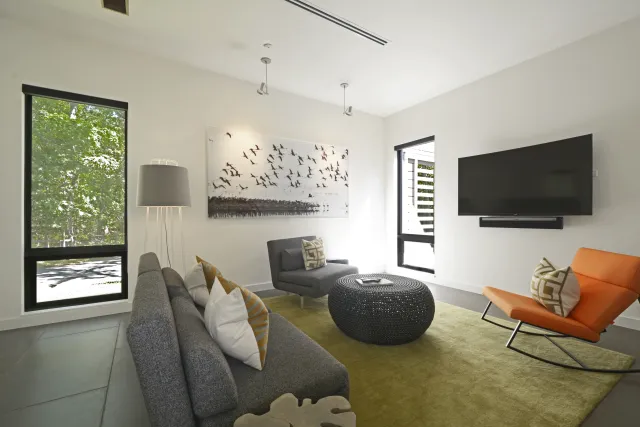 Modern living room with gray couch, orange chair, black ottoman, green rug, and wall-mounted TV.