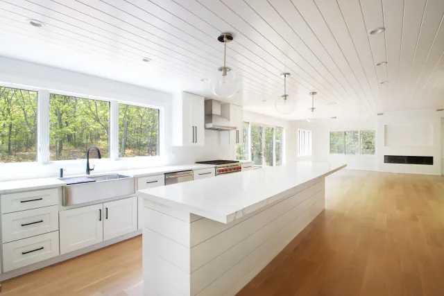 Bright modern open kitchen with white cabinetry, large island, wood floors, and large windows overlooking greenery.