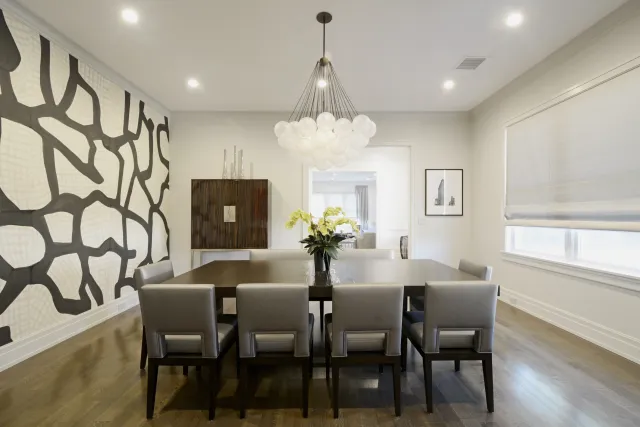 Modern dining room with large wooden table, eight grey chairs, abstract wall art, and a bubble chandelier.