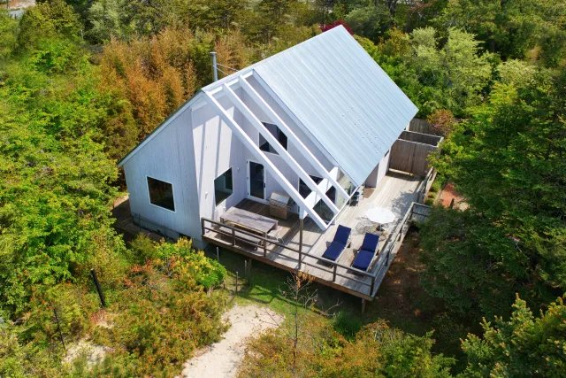Modern white cabin with large deck and seating surrounded by dense green forest in autumn setting