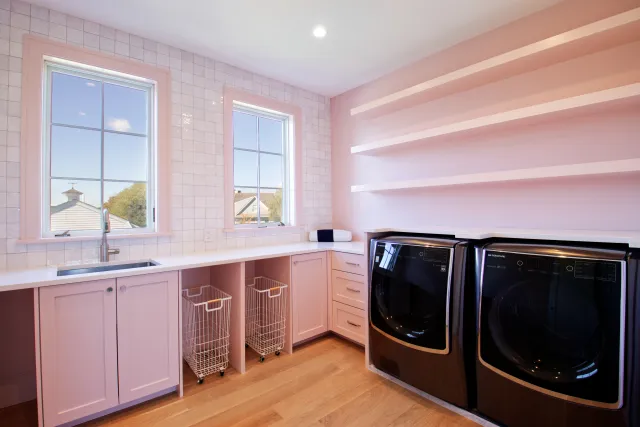 Bright modern laundry room with pink cabinets, black washer and dryer, white tiled walls, and wooden floor.