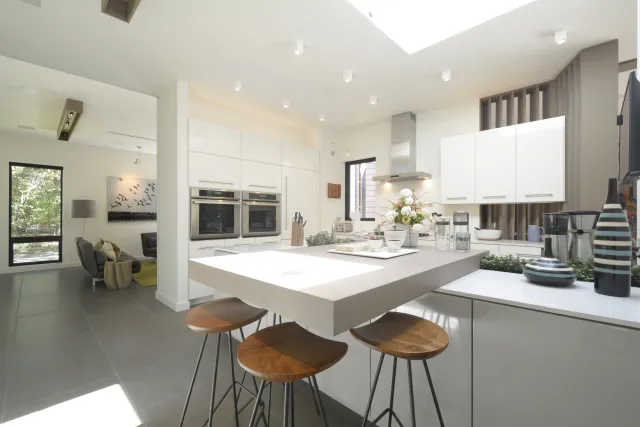 Bright modern kitchen with white cabinets, a large island with wooden stools, and sleek appliances under skylight.