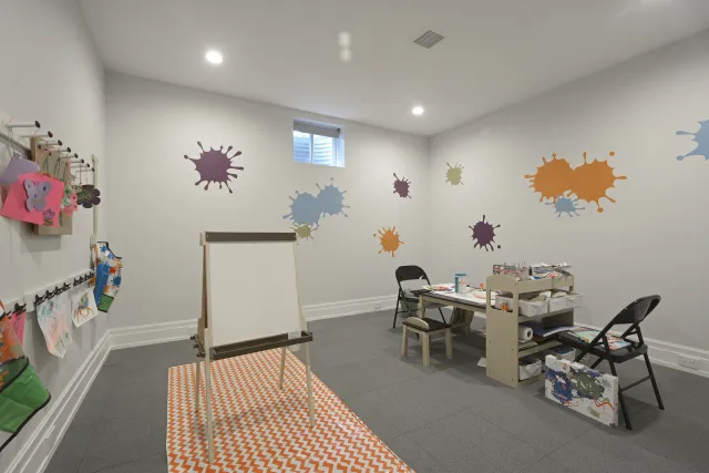 Bright art room with paint splatter wall decals, easel, tables, chairs, and kids' artwork hanging on wall hooks.