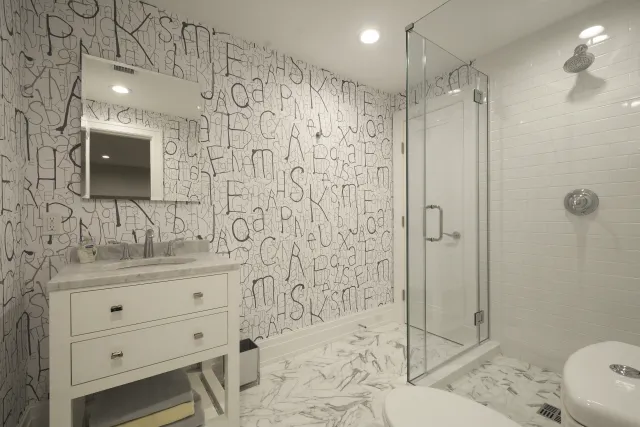 Modern bathroom with abstract letter wallpaper, white vanity, glass shower enclosure, and marble floors.