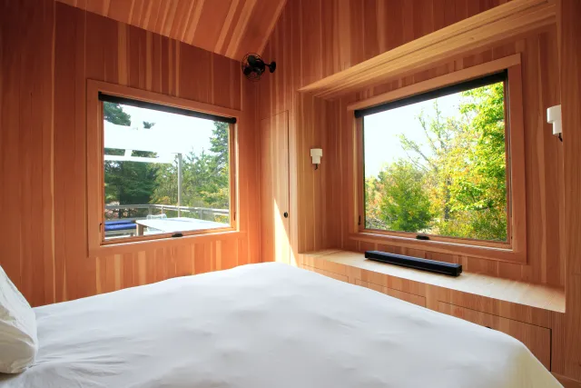 Cozy bedroom with wood-paneled walls and large windows showcasing green trees outside on a sunny day.