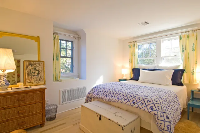 Bright bedroom with white walls, blue patterned bedding, wicker dresser, and large windows with patterned curtains.