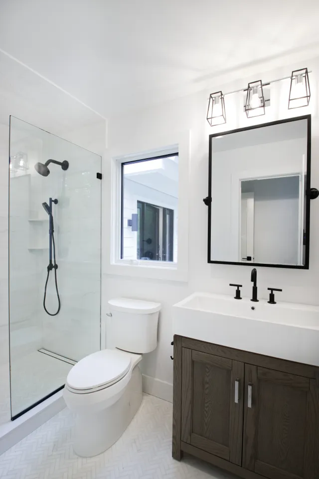 Modern white bathroom with glass shower, black fixtures, wooden vanity, large mirror, and bright lighting.