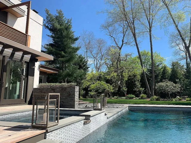 Modern backyard pool area with clear water, lanterns on the edge, and a lush green garden under a bright blue sky