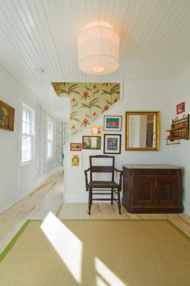 Bright vintage hallway featuring floral wallpaper, wooden chair, antique cabinet, gallery wall, and large area rug.