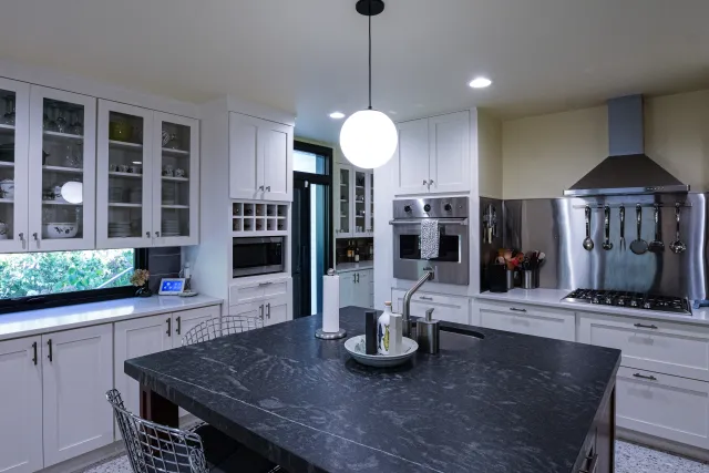 Modern kitchen with white cabinetry, black marble island countertop, stainless steel appliances, and hanging utensils.