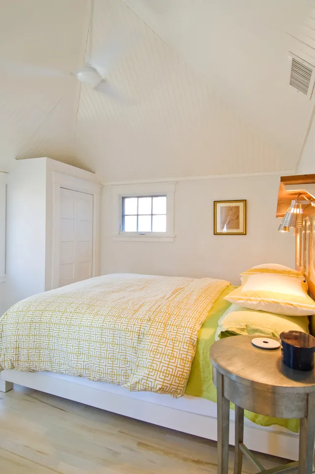 Bright bedroom with white walls, vaulted ceiling, yellow bedding, and round bedside table with blue candle.