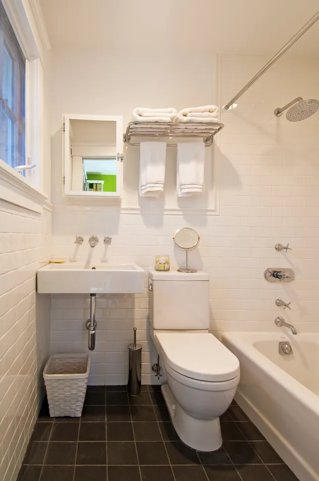 Modern bathroom with white tiles, a bathtub, toilet, sink, wall mirror, and neatly folded towels on a rack.