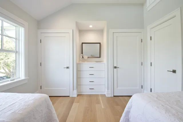 Bright bedroom with two white beds, wooden floor, white dresser with mirror, three closed doors, and large window.