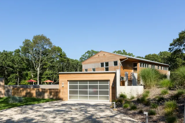 Modern wooden house with large windows and frosted garage door surrounded by greenery under a clear blue sky