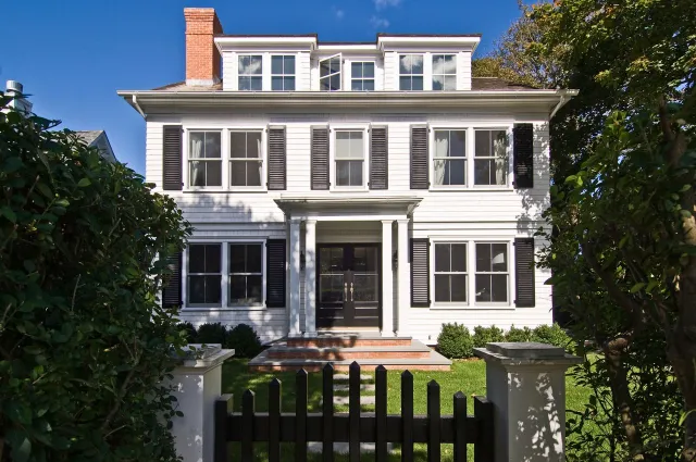 White colonial-style house with black shutters, brick chimney, and a black picket fence surrounded by greenery.