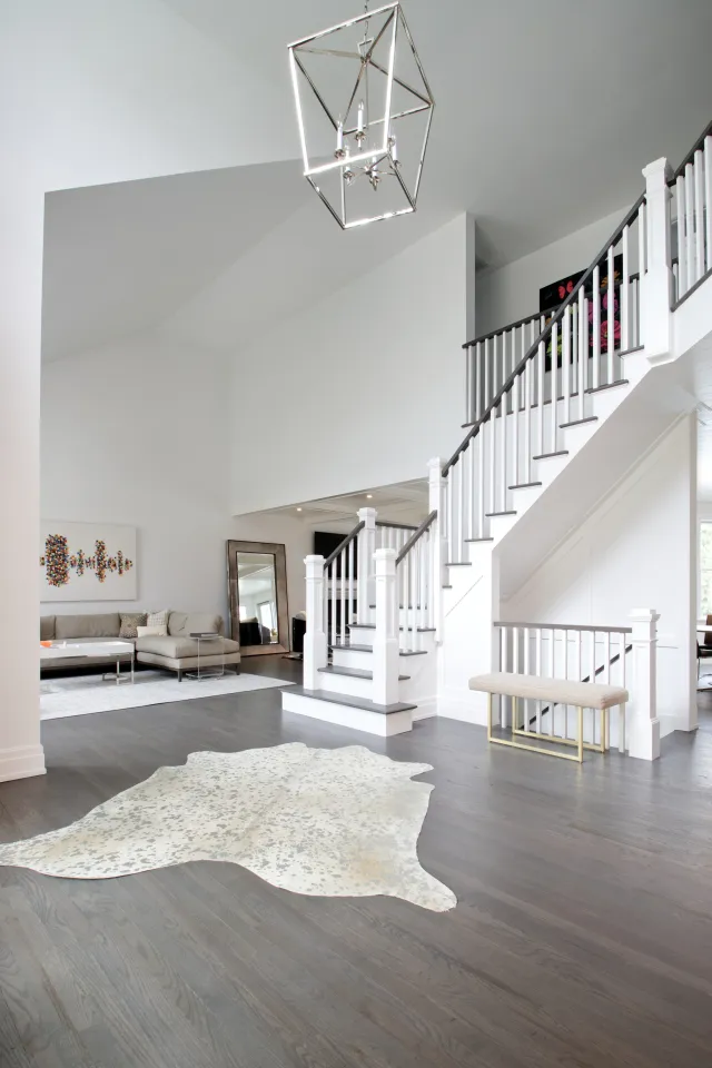Spacious modern living room with white walls, dark hardwood floors, cowhide rug, staircase, and beige sectional sofa.