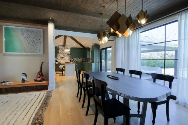 Modern dining room with black chairs, wooden floor, geometric pendant lights, and open kitchen with green cabinets.