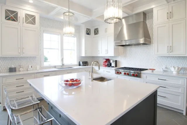 Bright modern kitchen with white cabinetry, large island, clear stools, and stainless steel appliances under chandelier lighting.